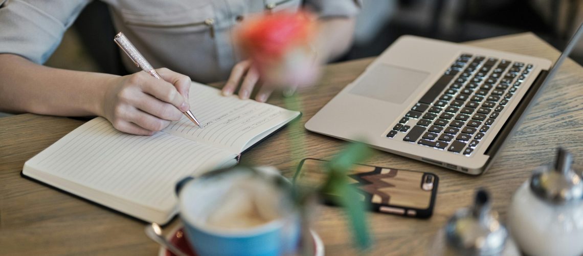 Woman writing in a notebook with a laptop and coffee cup on a desk. Ideal for workspace inspiration.