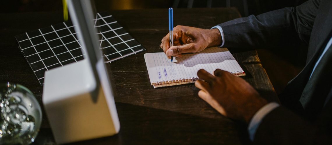 A businessman in a suit writing notes on a notebook at a desk, suggesting a professional office environment.