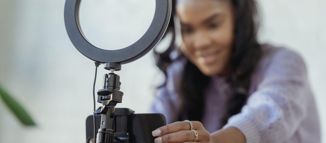 Cheerful young African American female blogger in stylish sweater smiling while setting up camera of smartphone attached to tripod with ring light before recording vlog