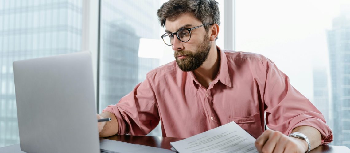 A businessman in an office reviews documents and data on a laptop, concentrating on financial analysis.